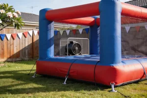 A red and blue jumping castle pegged down with straps on a backyard lawn, its blower in a guarded cage and bunting along the fence