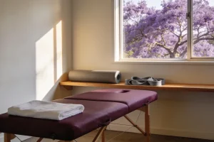 An empty treatment room with a padded table and folded towel, a foam roller and resistance bands on a shelf and a flowering jacaranda outside
